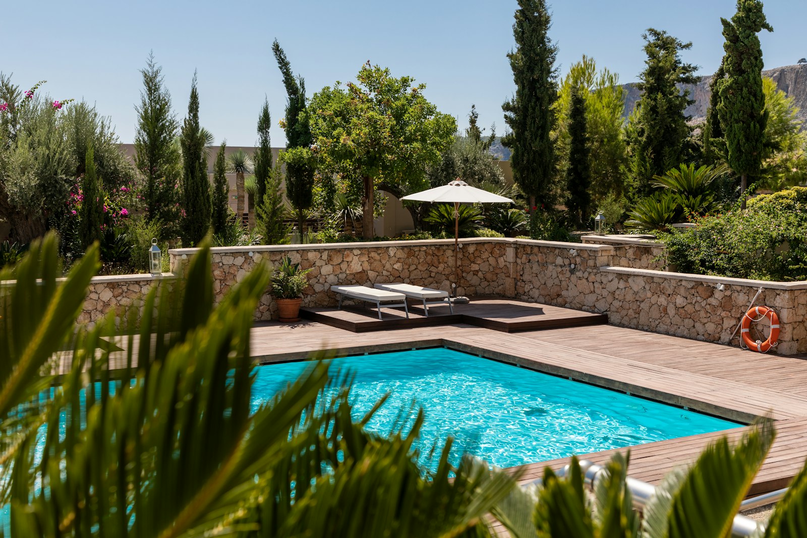 A landscaped residential pool with sparkling blue water, wood deck, sun loungers, and a shade umbrella